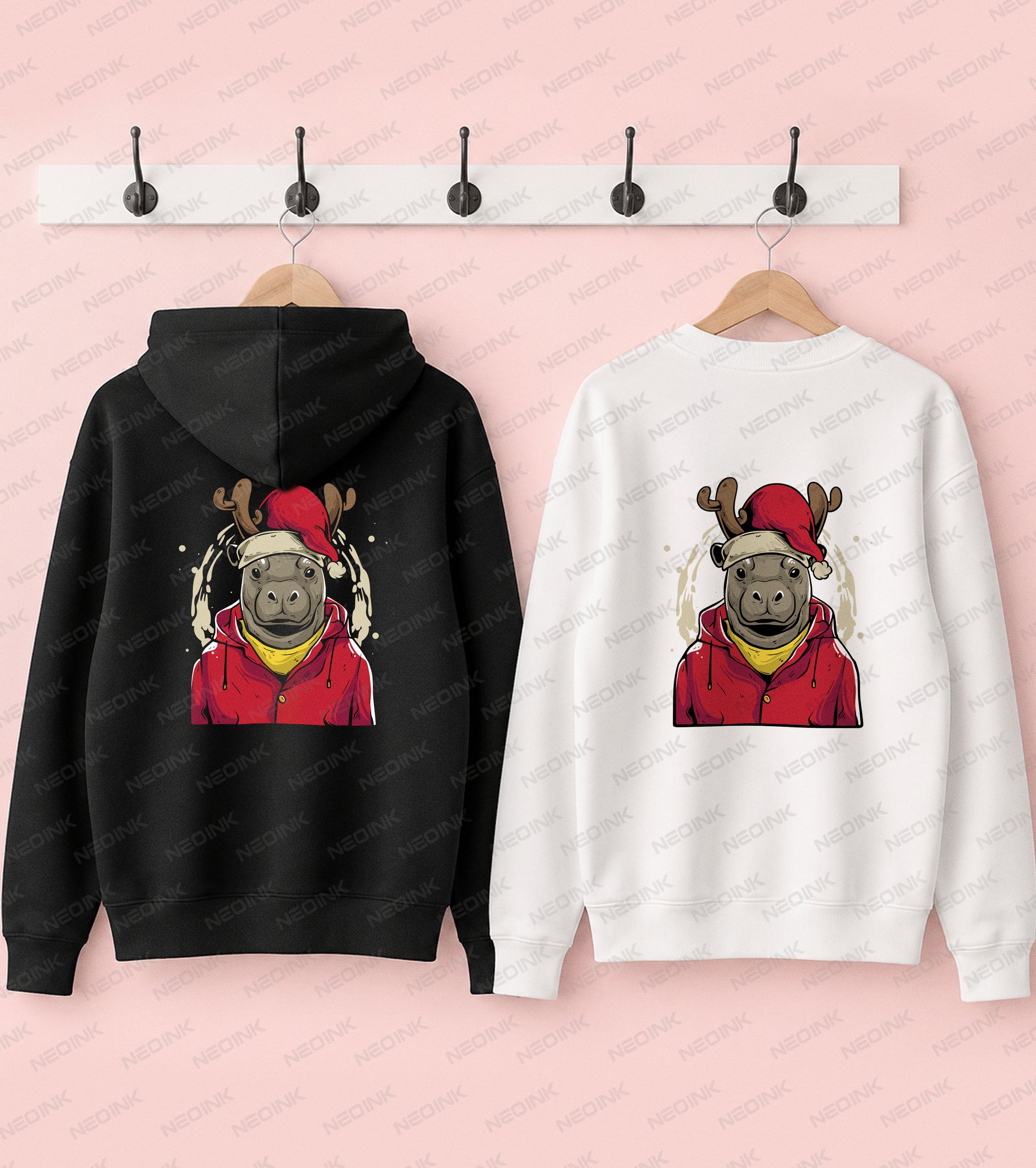 Two hoodies shown side by side on wooden hangers against a pink background, with a white coat rack above them. The left hoodie is black and the right hoodie is white, both featuring a cartoon reindeer wearing a red Santa hat and red jacket with a yellow scarf in the chest graphic.